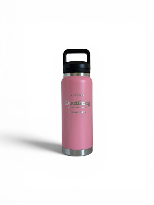 Pink stainless steel insulated water bottle with a black screw-on lid and built-in carry handle. The front features a laser engraved TiredGang logo with the phrases “ESTD MMXXV” and “RUNNING ON 1%.” Pink finish with stainless steel accents at the top and bottom. Product shown centered on a white background.